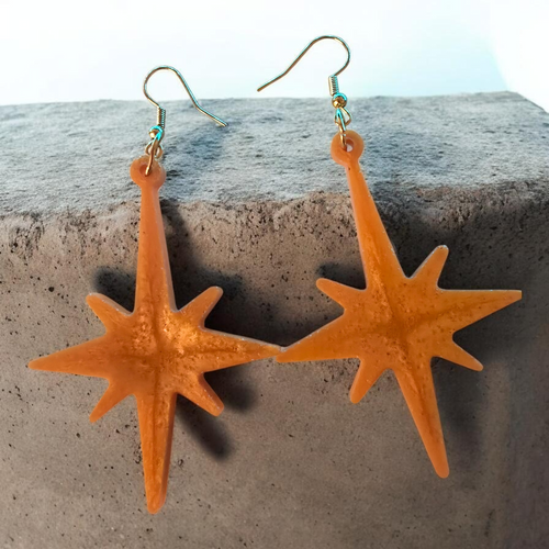 Love For Resin orange starburst resin dangle earrings with gold-tone hooks resting on a grey stone surface